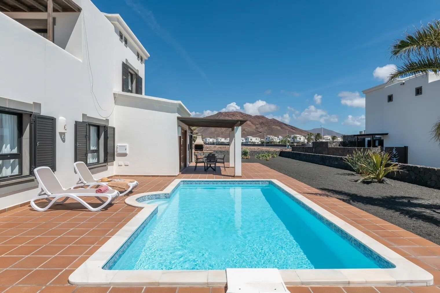 A rectangular swimming pool with a tiled deck, two lounge chairs, and a covered seating area with a table and chairs.