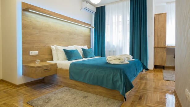 Una habitación de hotel moderna con cabecera de madera, cama con ropa de cama color turquesa, mesita de noche, armario y piso de madera.
