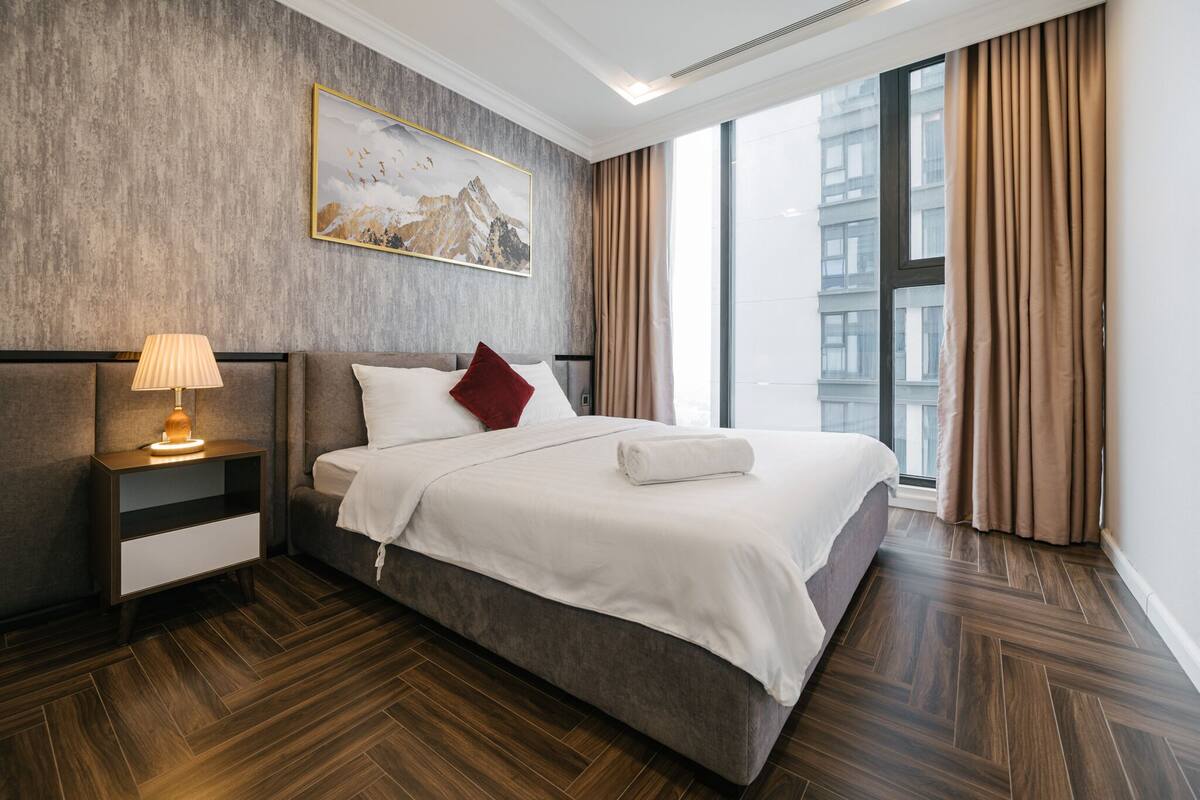A modern hotel room with a large bed, a nightstand with a lamp, a painting on the wall, and a view of the city through large windows.