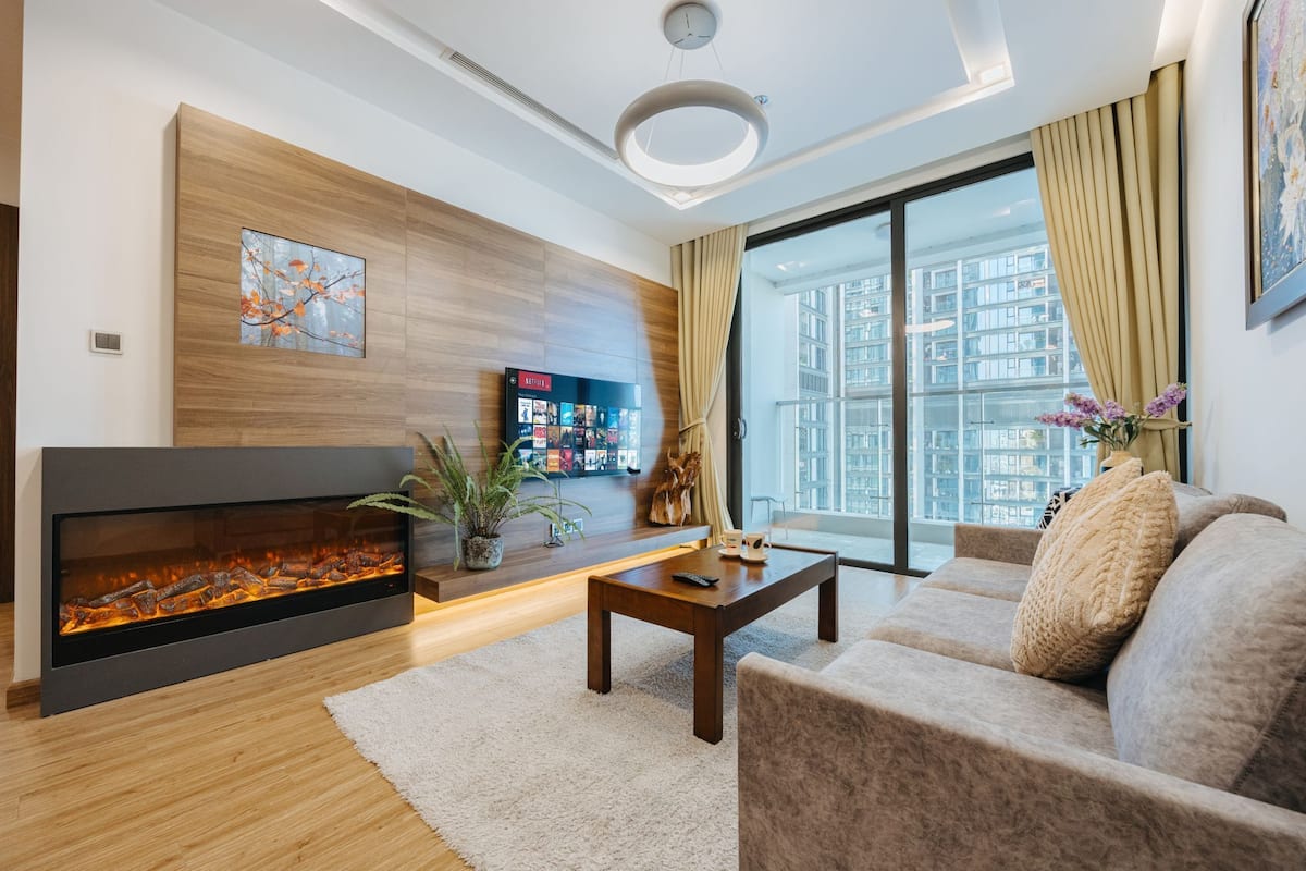 A modern living room with a fireplace, a sofa, a coffee table, and a large window offering a city view.