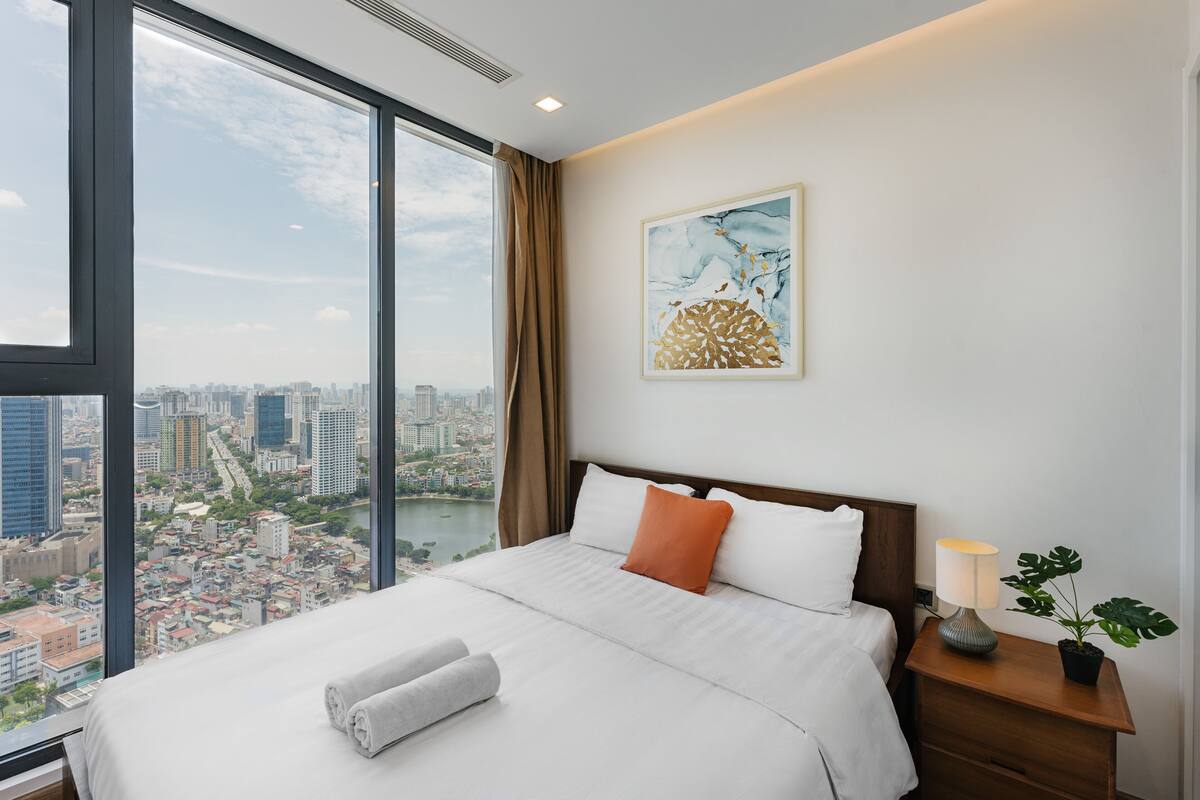 A hotel room with a large window, a bed with white linens, an orange pillow, a nightstand with a lamp, and a city view.