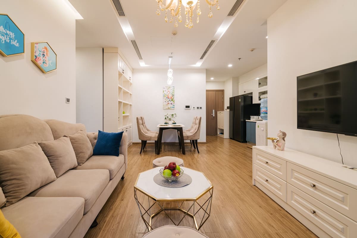 A modern living room with a beige sofa, a coffee table with fruit, a dining area with chairs, and a TV on a cabinet.