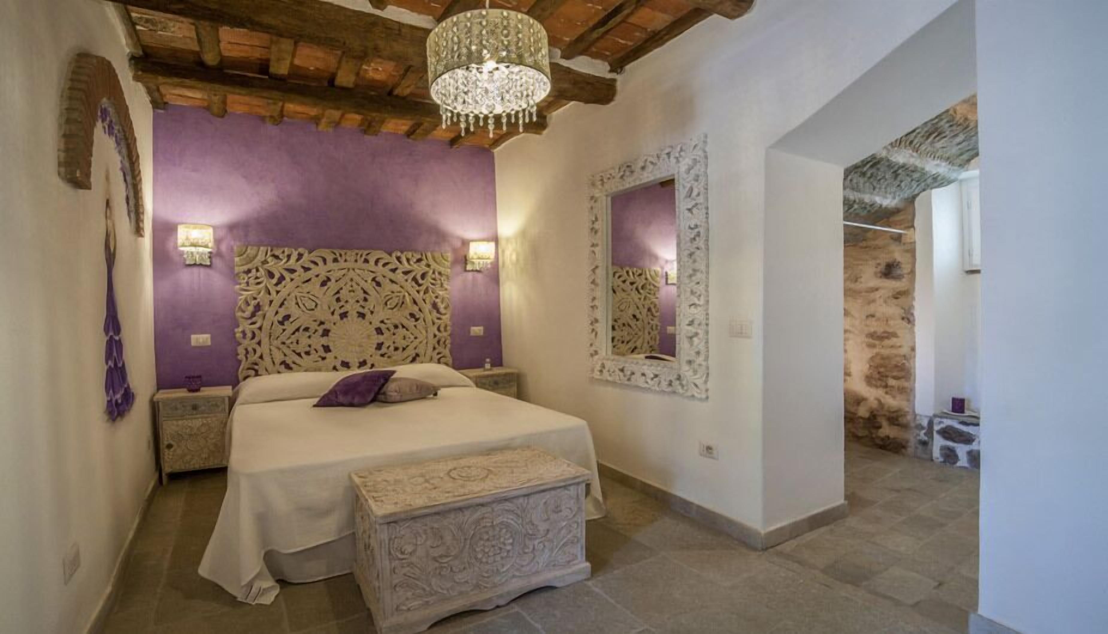 A bedroom with a large bed, a decorative headboard, two wall-mounted lights, a mirror, and a stone bench.