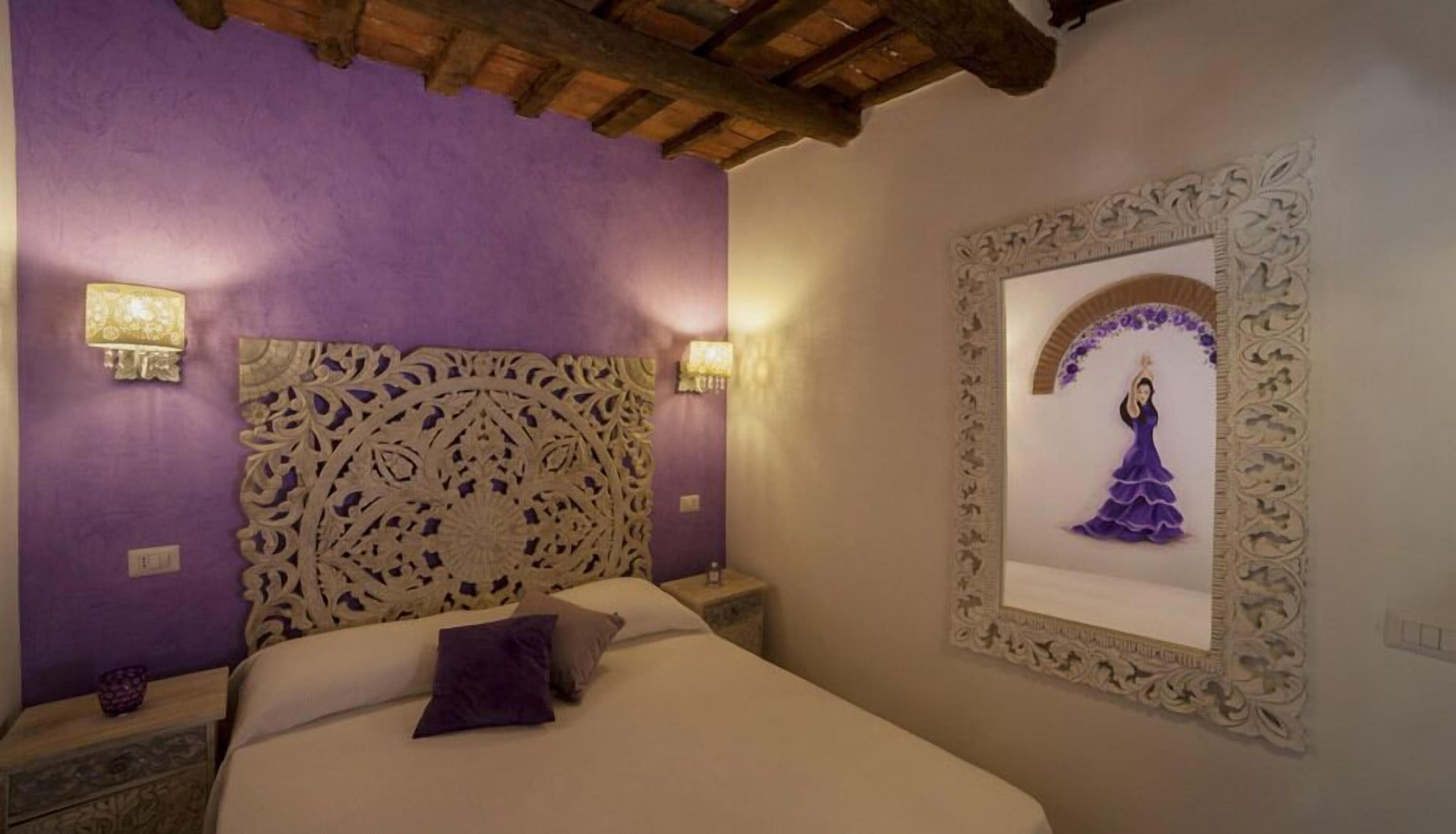A bedroom with a bed, purple walls, a decorative headboard, and a framed artwork on the wall.