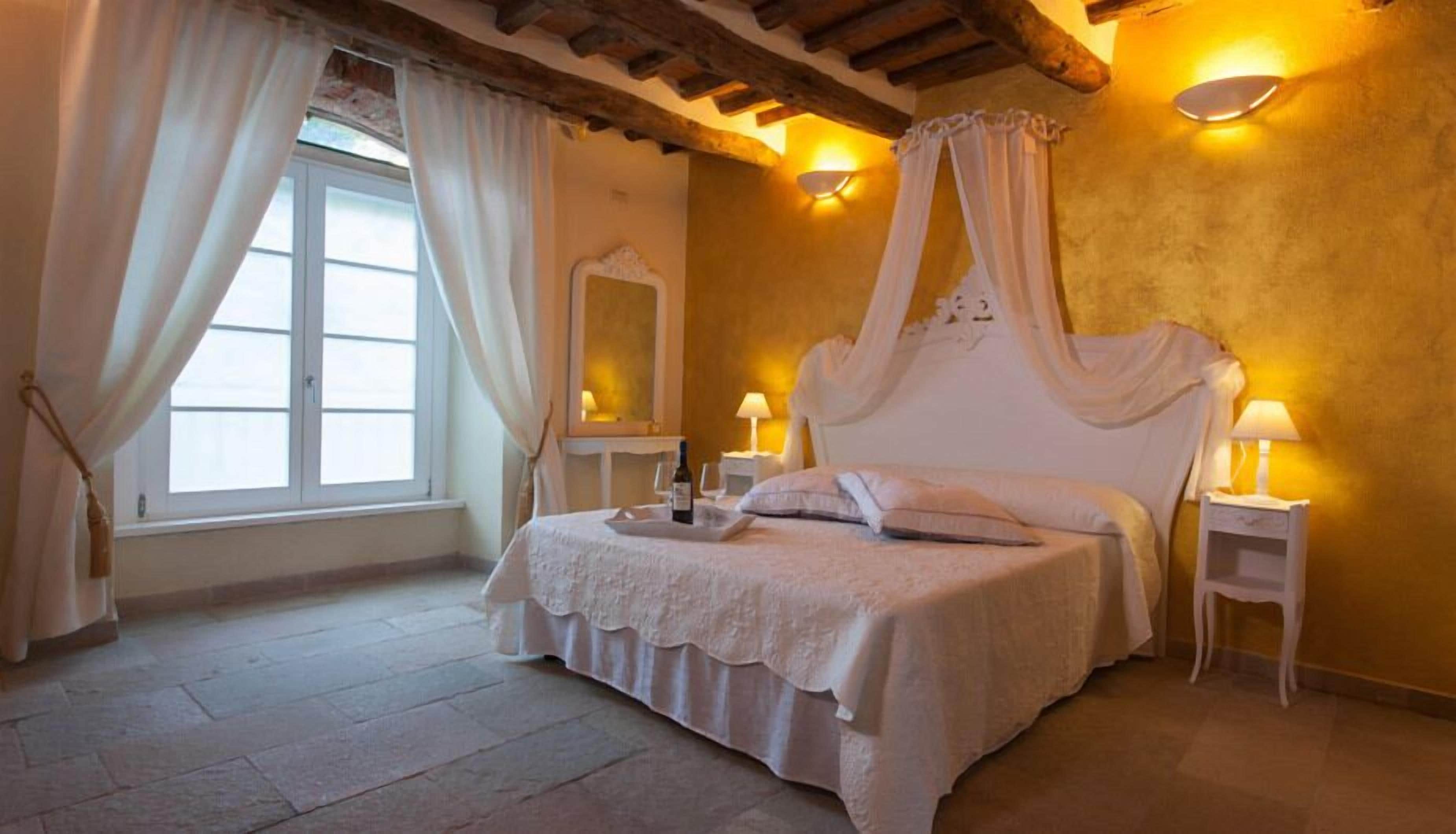 Een slaapkamer met een hemelbed, twee nachtkastjes, een raam met gordijnen en wandlampen.