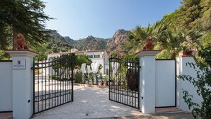A gated entrance to a property with a white building, two lion statues on pillars, and a mountainous backdrop. - Bed & Breakfast Il Casale di Monserrato (Porto Azzurro)