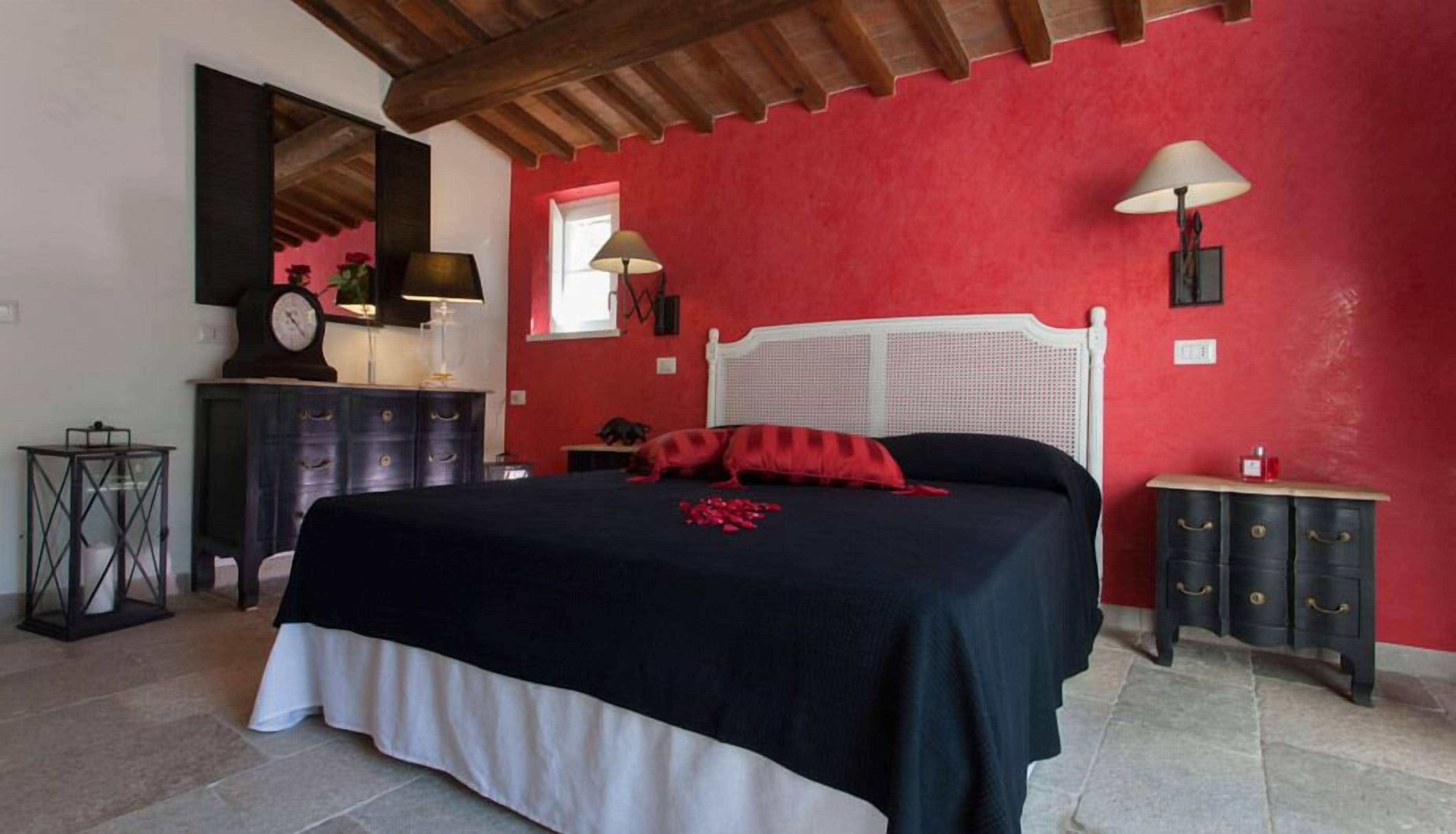 A bedroom with a red wall, a white headboard, a black bedspread, and a red throw pillow. There is a wooden dresser with drawers and a lamp on it.