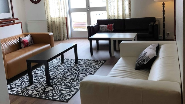 A living room with two sofas, a coffee table, and a dining table. There is a large window with curtains, an air conditioning unit, and a mirror on the wall.