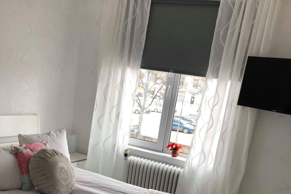 Une chambre à coucher avec un lit, des rideaux blancs, un radiateur et une fenêtre donnant sur la rue.