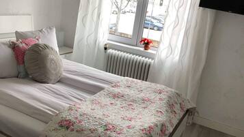 A bedroom with a bed, white curtains, a radiator, and a window with a view of the street.