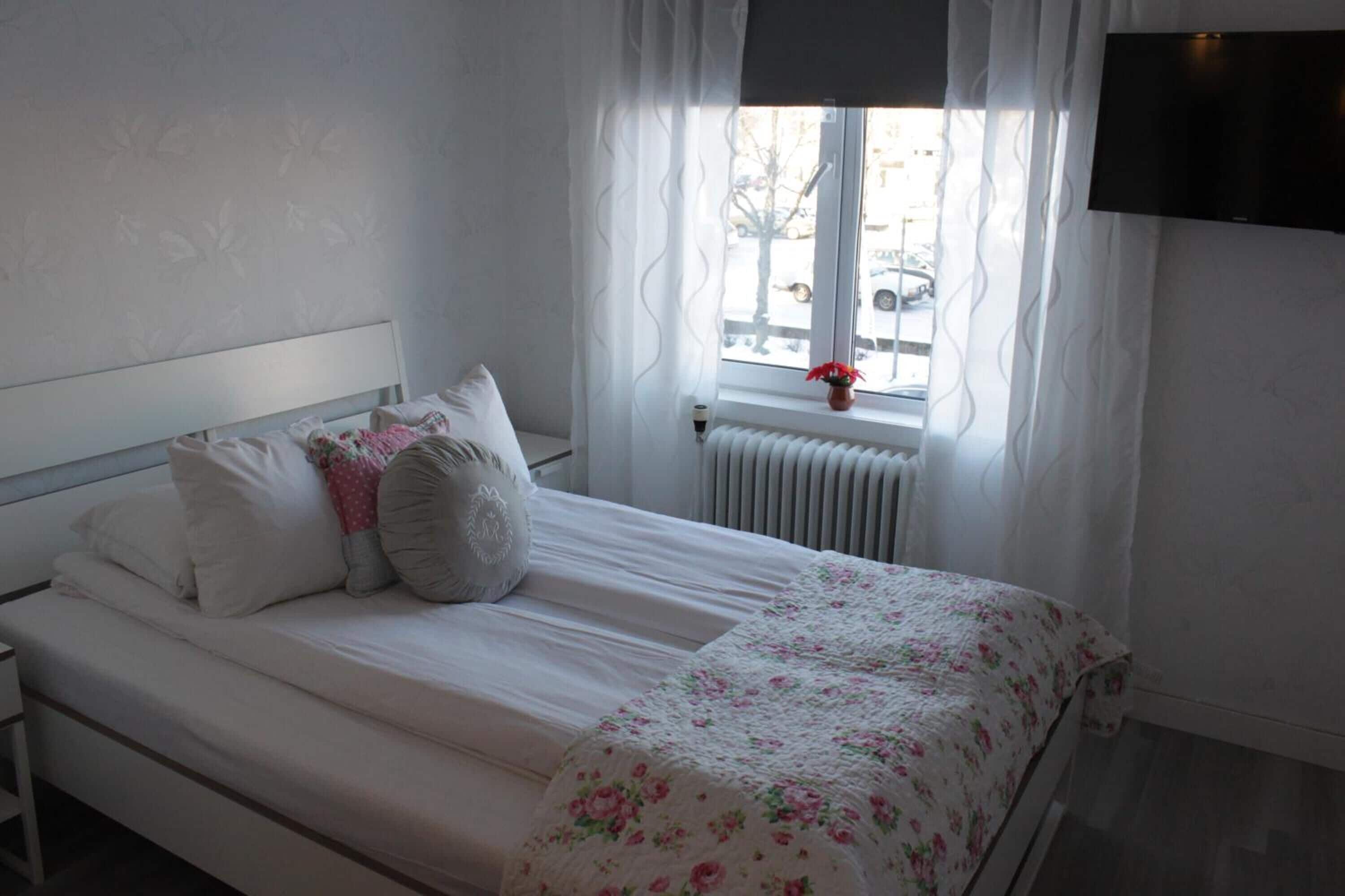 A bedroom with a bed, white bedding, a window with curtains, a radiator, and a TV mounted on the wall.