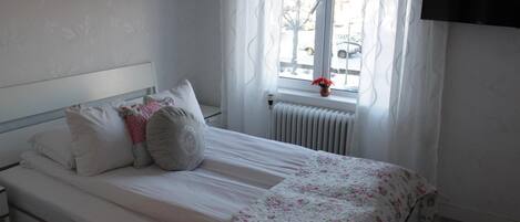 A bedroom with a bed, white bedding, a window with curtains, a radiator, and a TV mounted on the wall.