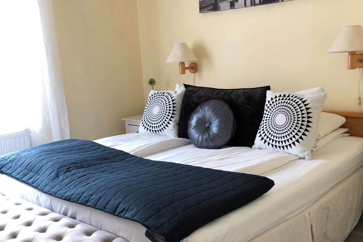 Un lit bien fait, recouvert d’une courtepointe bleu foncé, agrémenté de deux coussins décoratifs et surmonté d’un banc placé au pied du lit.