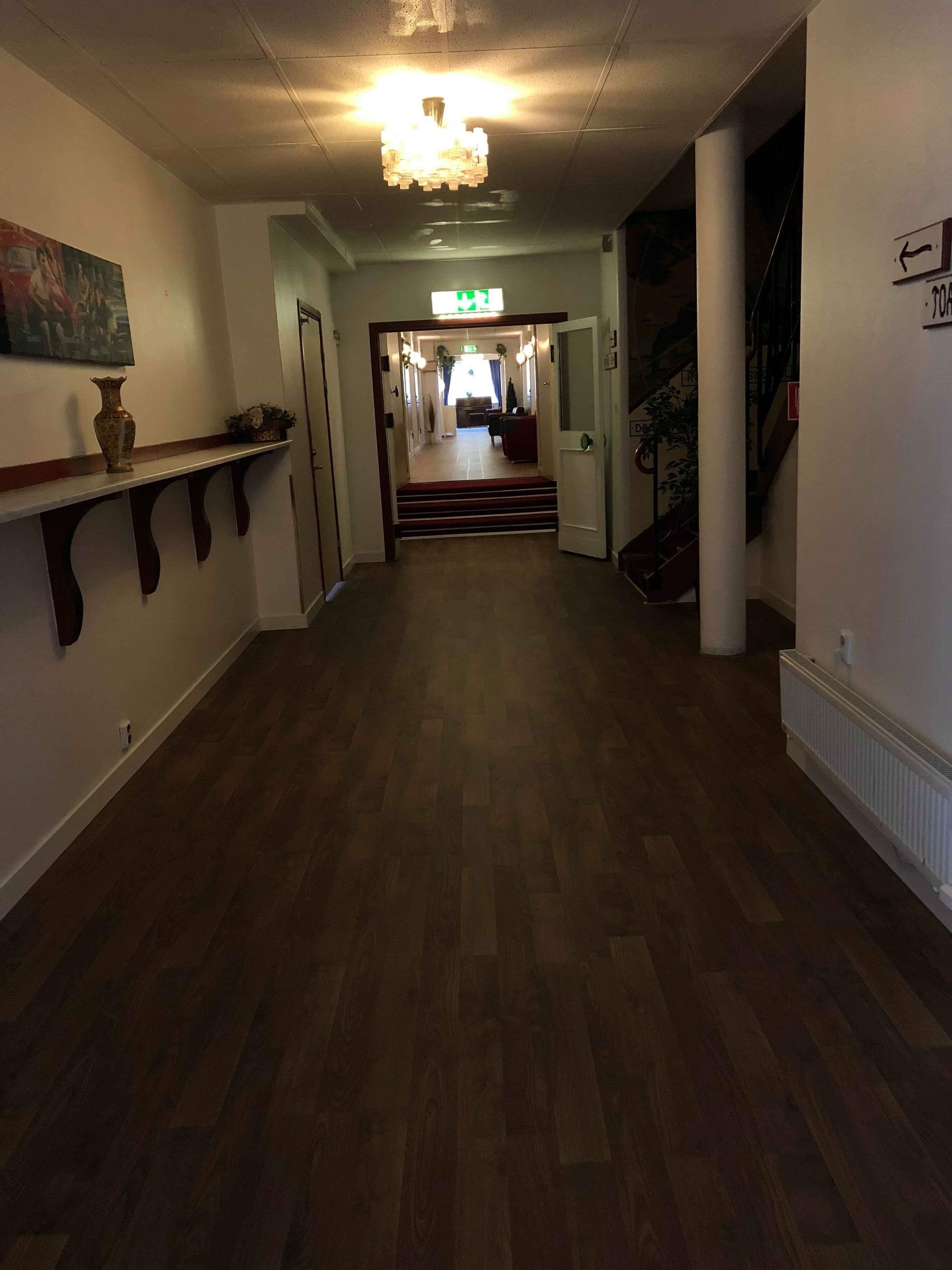 Un corridoio d'albergo con pavimento in legno, un lampadario, un quadro appeso alla parete e una scala che conduce ad un'altra zona.