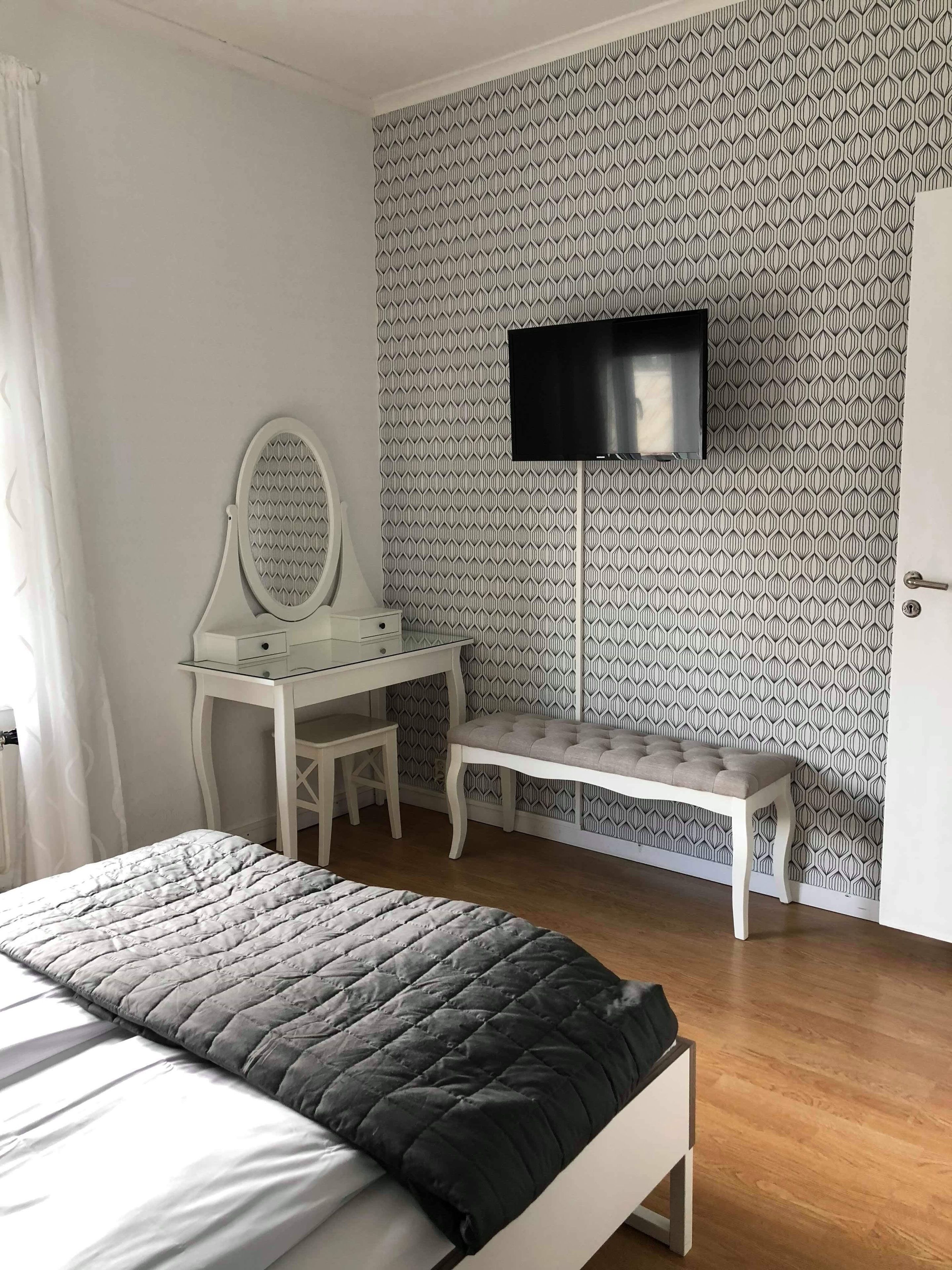 Un dormitorio con cama, escritorio, silla, banco y televisor montado en la pared.