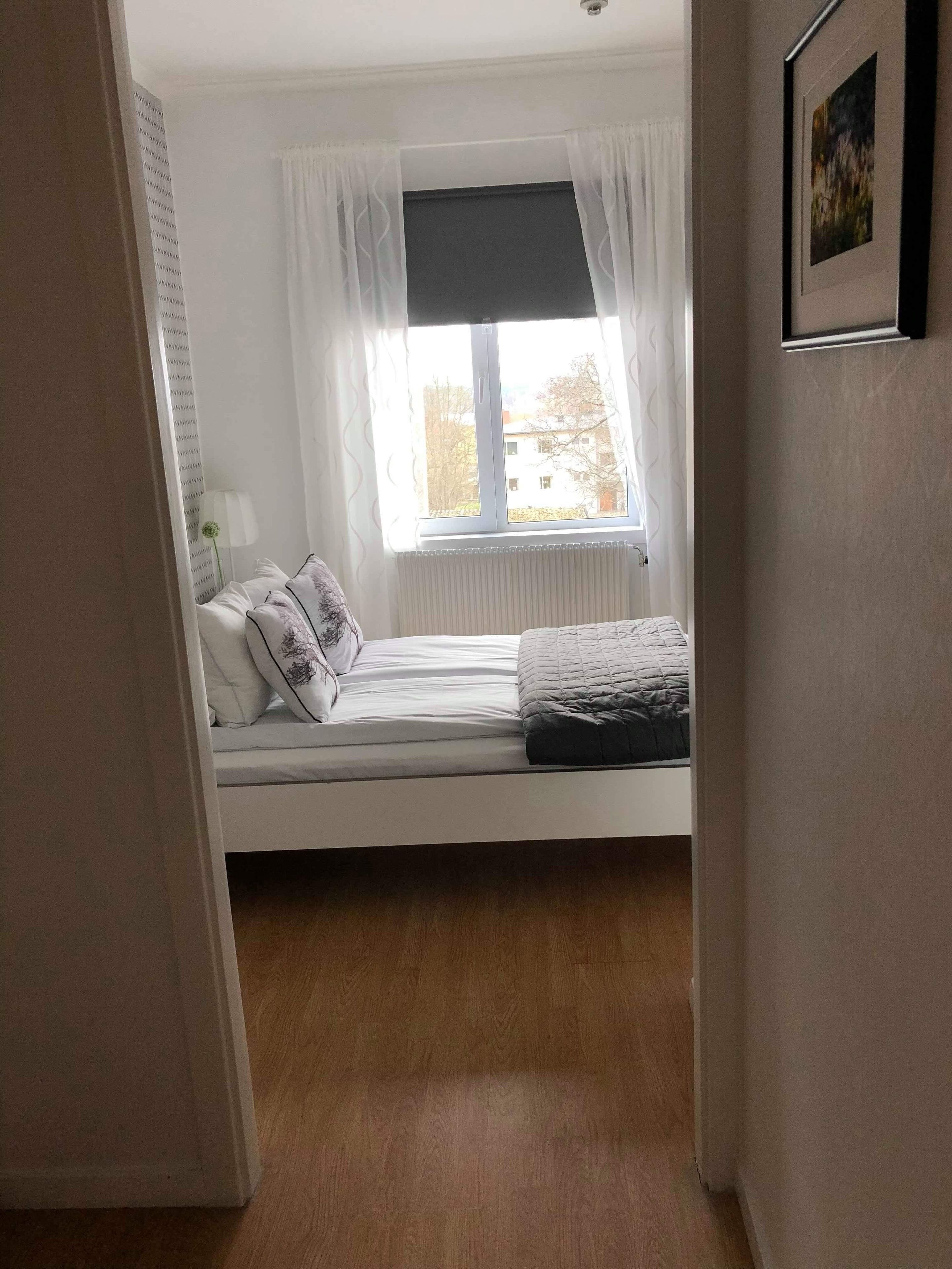 Un dormitorio con cama, cortinas blancas, ventana y radiador.