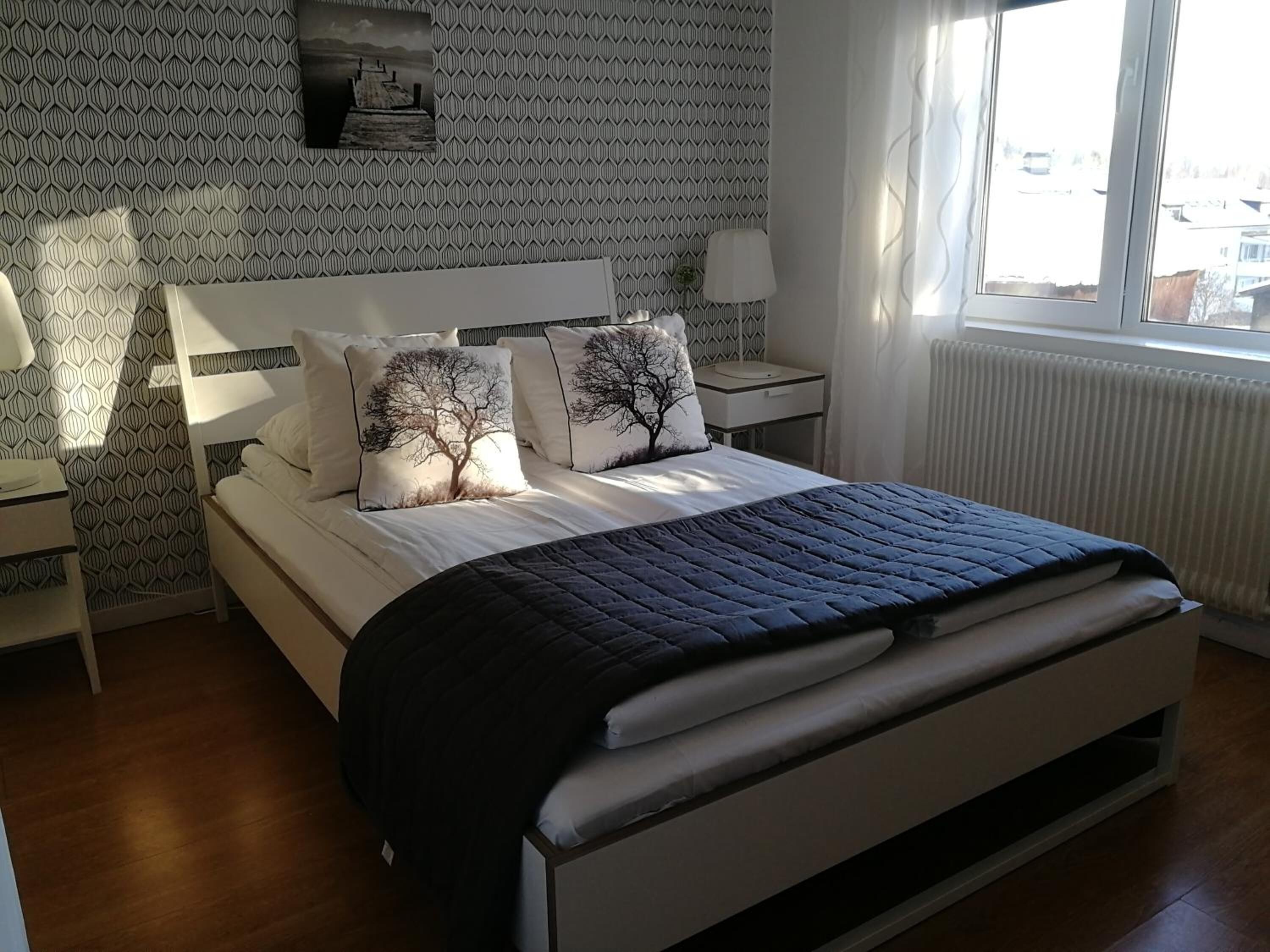 A bedroom with a bed, two nightstands, a window, and patterned wallpaper.