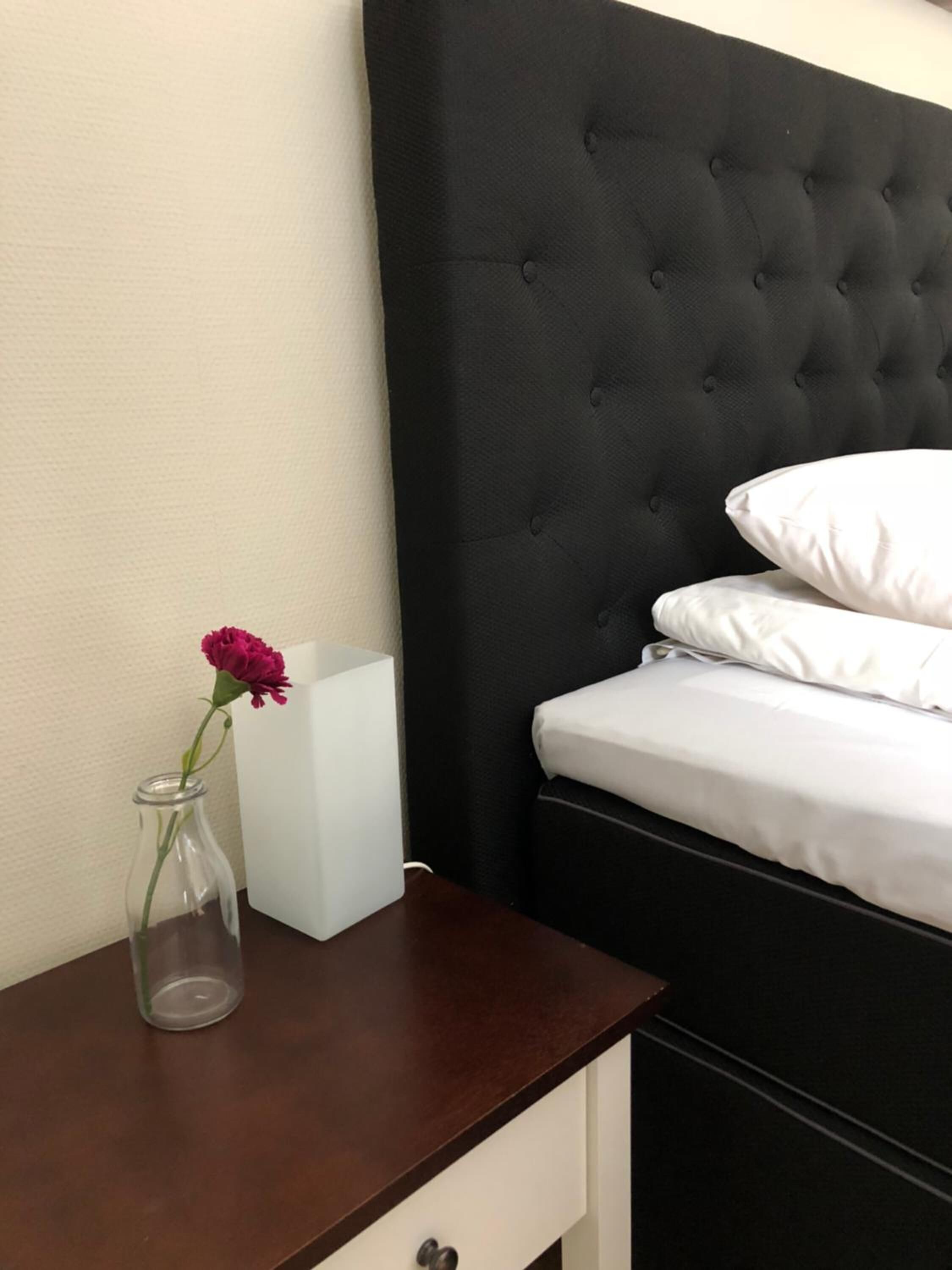 Un dormitorio con cabecera tapizada en tonos oscuros, ropa de cama blanca, una mesita de noche con un florero y una unidad de mesita de noche blanca.