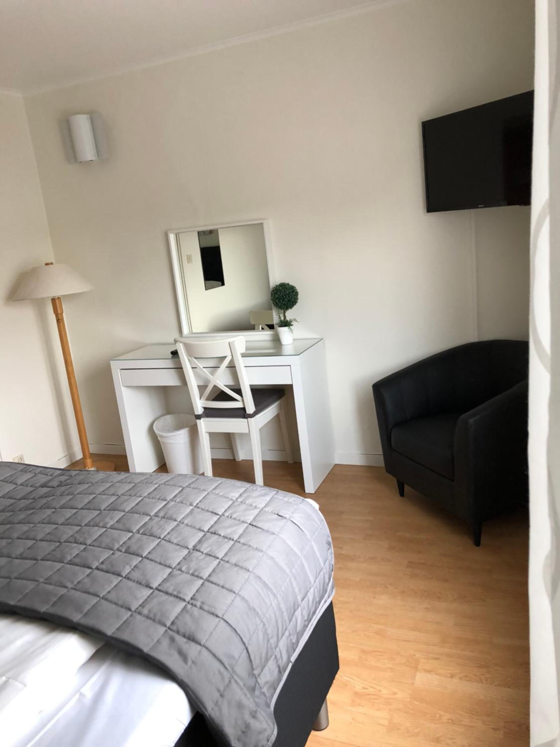 Un dormitorio con cama, escritorio, silla, sofá negro, planta pequeña en maceta, espejo y televisor montado en la pared.