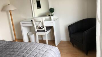 Un dormitorio con cama, escritorio, silla, sofá negro, planta pequeña en maceta, espejo y televisor montado en la pared.