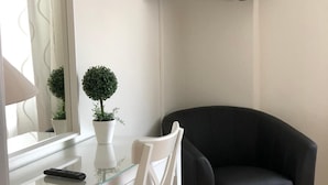 A modern room with a glass desk, a white chair, a black sofa, a mounted TV, and two potted plants.