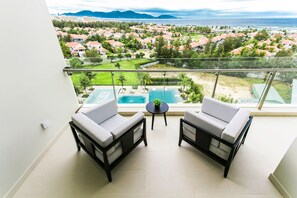 A balcony with two white sofas and a small table, overlooking a pool and a residential area with red-roofed houses.