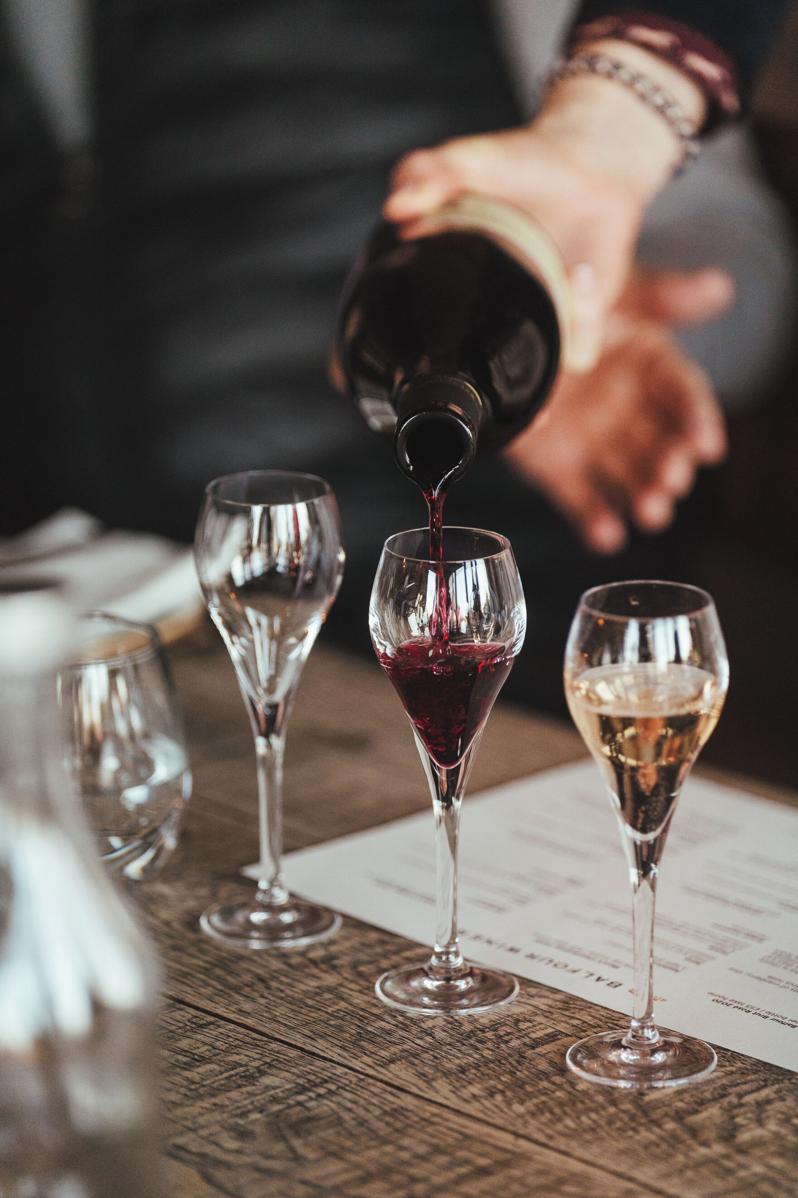 A person pouring wine into a glass, with other glasses and a menu on a wooden table.