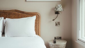 A neatly made bed with white linens, a woven headboard, a bedside lamp, a small round table with a remote control, and a framed picture on the wall. - Winemaker's Pub (Cranbrook)