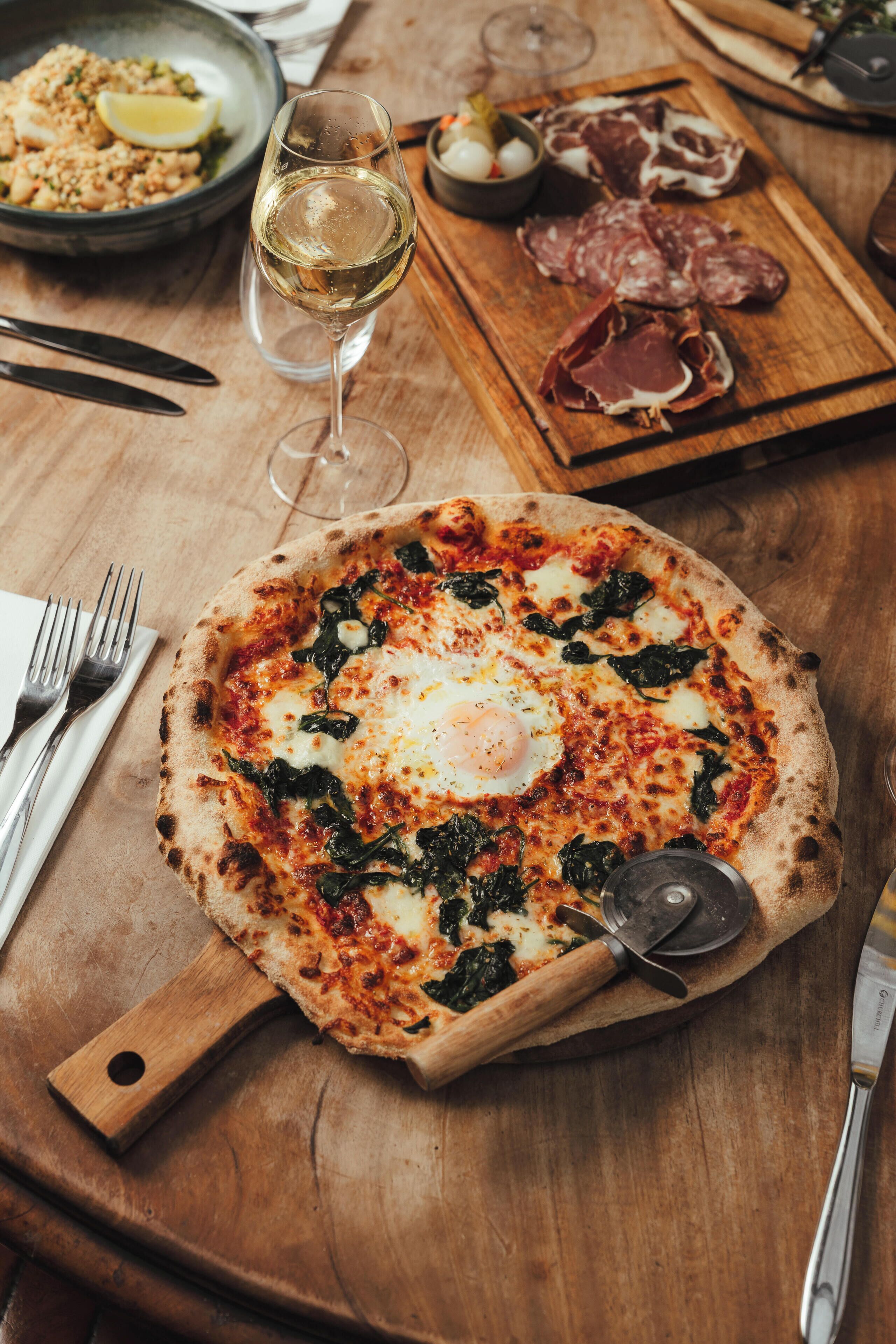 Pizza mit Spinat und Ei auf einem Holzbalken, ein Glas Weißwein und eine Platte mit Wurstwaren.