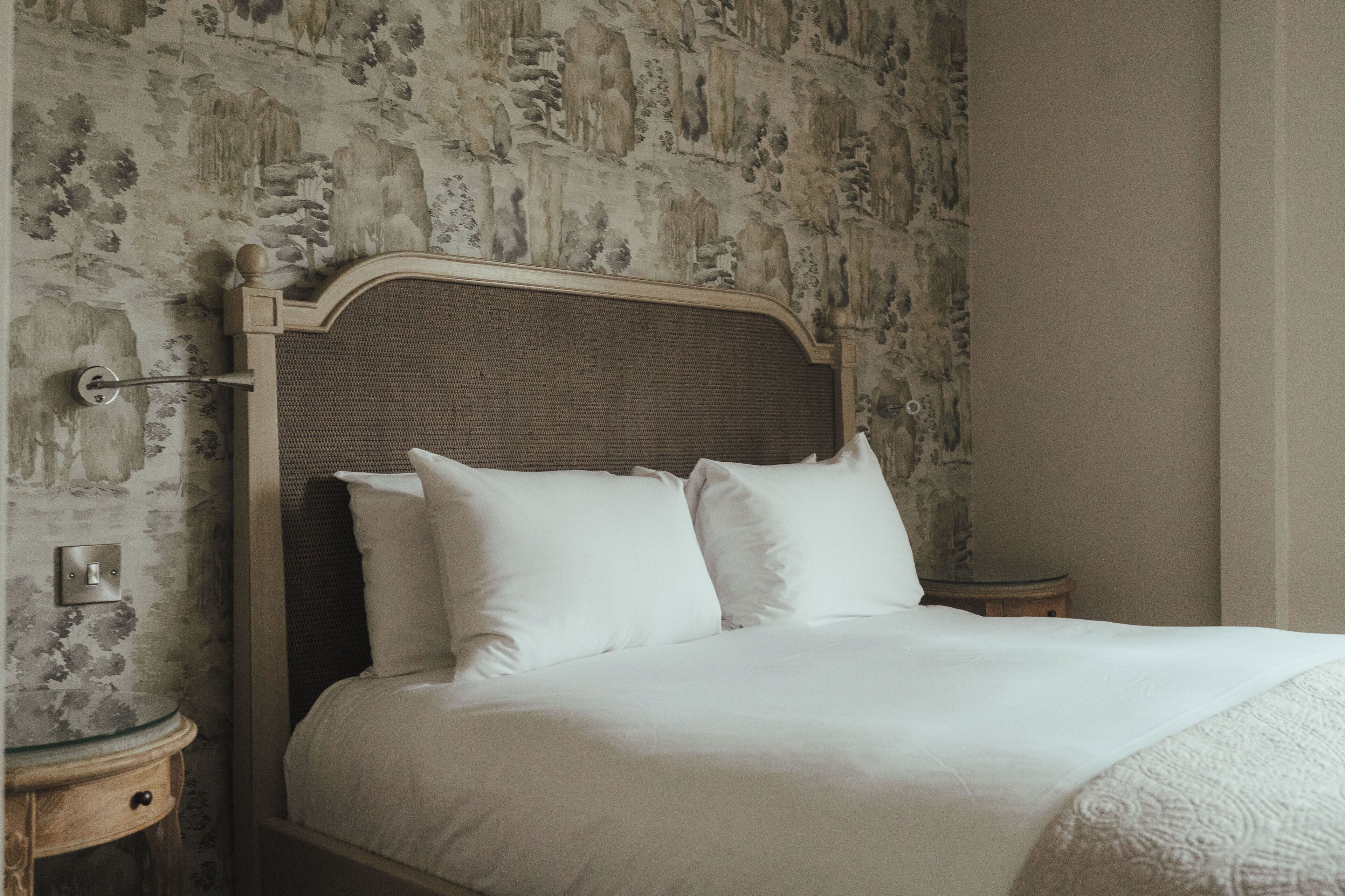 Una cama con cabecera trenzada, ropa de cama blanca y una pared con estampado floral.