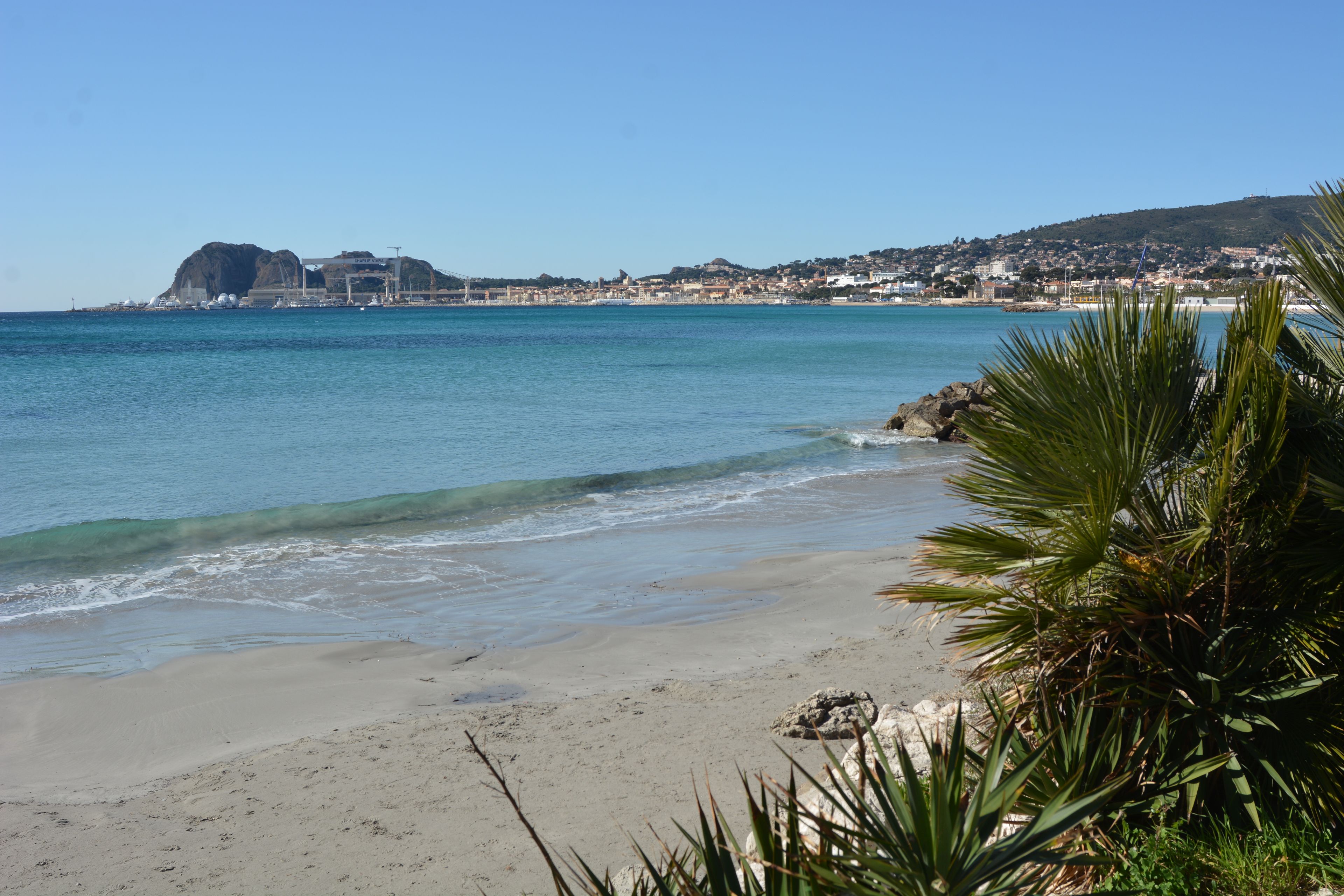 Una spiaggia con acque limpide, un litorale roccioso e una vista in lontananza di una cittadina con edifici e una collina.