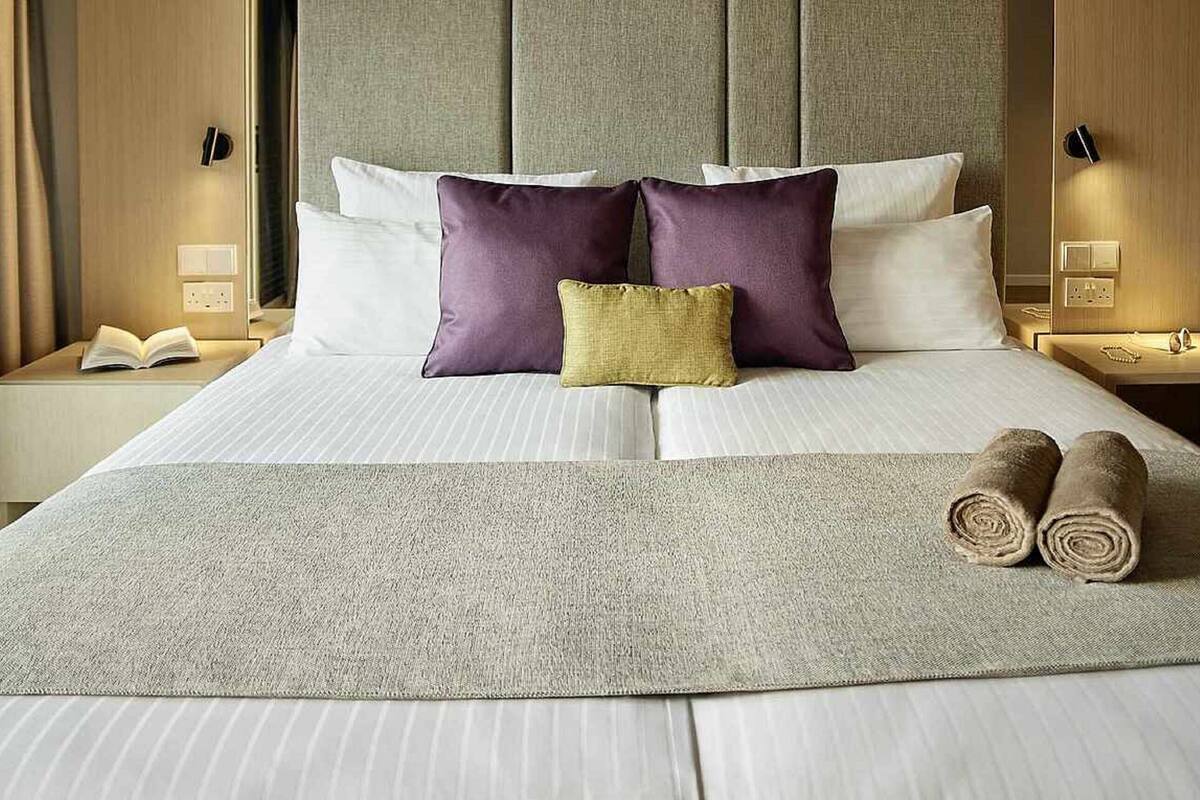 A neatly made bed with white linens, two purple pillows, a small yellow pillow, and a beige throw blanket. There are two bedside tables with lamps and a book on one of them.