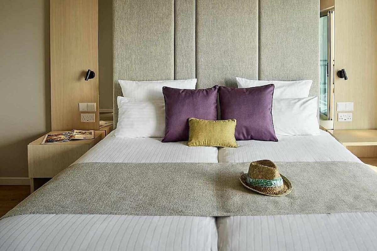 A neatly made bed with white and purple pillows, a woven throw blanket, and a straw hat on the bed.