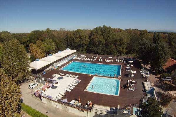 Una gran piscina al aire libre con múltiples carriles, rodeada de sillas de descanso y sombrillas, con árboles y un cielo despejado de fondo.