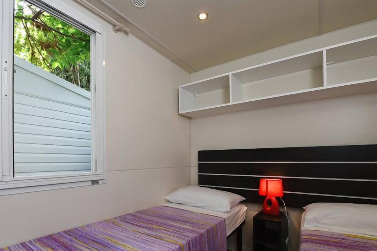 A room with two single beds, a window with blinds, a red lamp on a nightstand, and built-in shelving above the beds.
