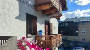 A building with a wooden balcony, a flower bed with pink and white flowers, and a clear blue sky with clouds. - Hotel Aquila (Livigno)