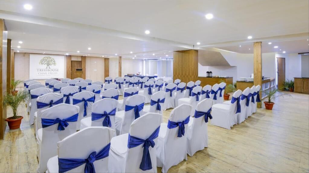Une salle de conférence avec des rangées de chaises blanches ornées de rubans bleus, un pupitre en bois et une enseigne indiquant « Treehouse Chalet Villas ».