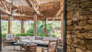 A cozy outdoor seating area with a sofa, rocking chair, and tables, surrounded by natural stone walls and a thatched roof. - Saseka Tented Camp (Hoedspruit)