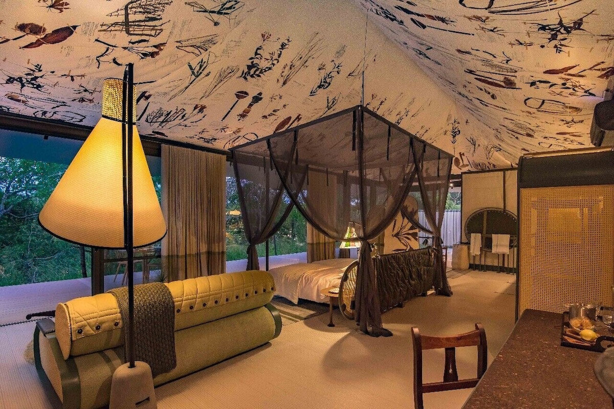A room with a canopy bed, a sofa, a dining table, and a large window with a view of greenery.