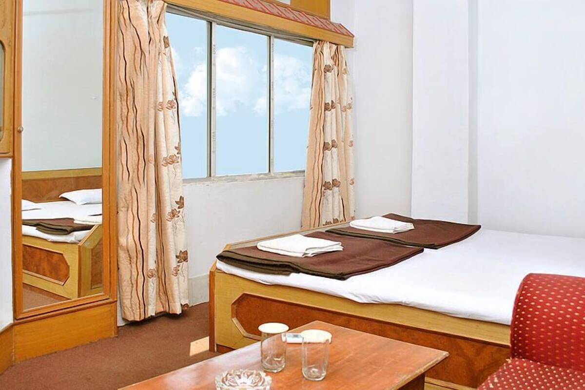 A hotel room with a bed, a wooden table with two glasses, a red chair, and a window with curtains.