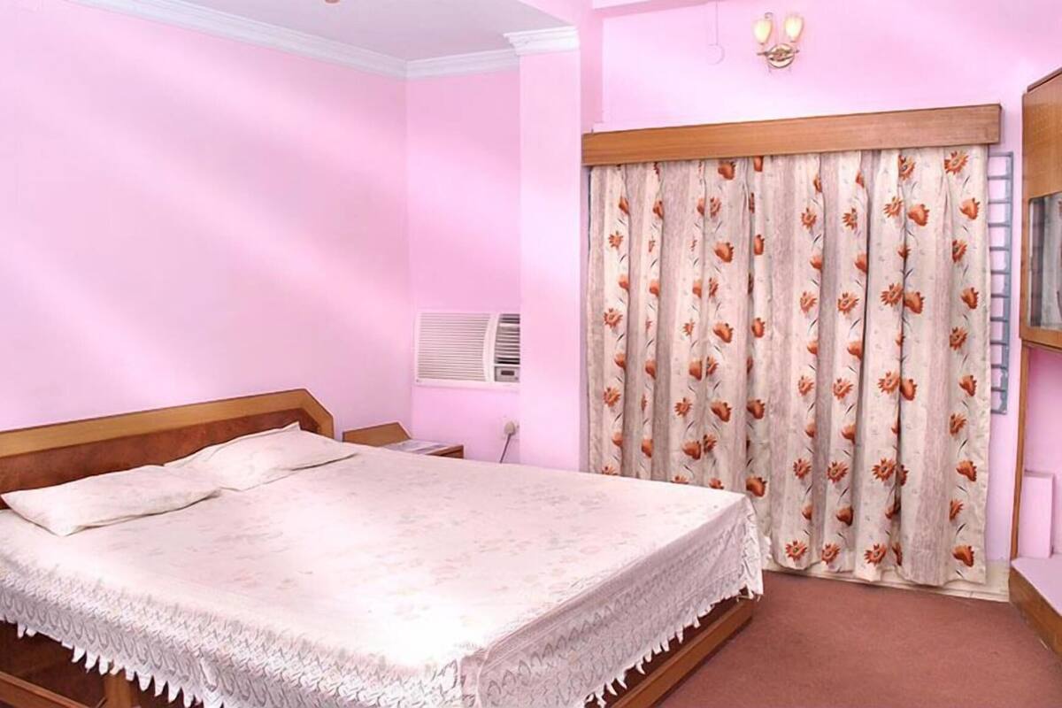 A bedroom with a bed, a nightstand, a wall-mounted air conditioner, and a window with floral curtains.