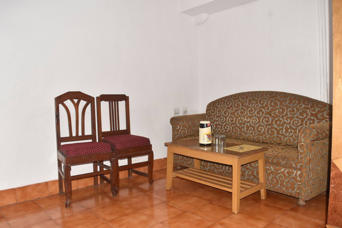 A room with a patterned sofa, two wooden chairs, and a small wooden table with a coffee maker.