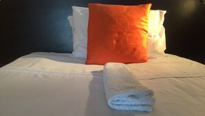 Een net opgemaakt bed met witte lakens, een opgevouwen witte handdoek en twee kussens (een oranje en een wit).