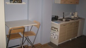 Uma kitchenette compacta com pia, fogão e armários. Há uma pequena mesa com duas cadeiras e uma estante com um quadro.