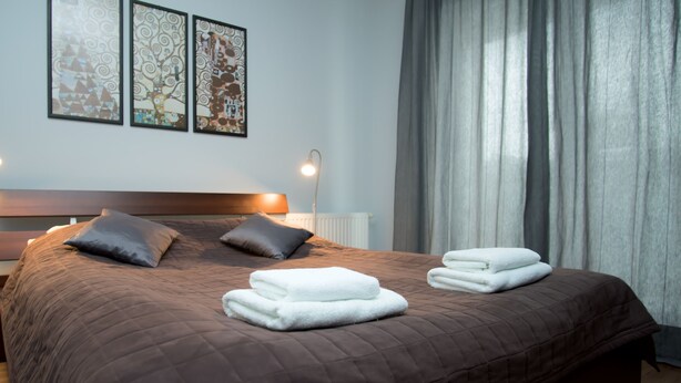 Ein ordentlich bezogenes Bett mit einer braunen Steude, zwei Kissen und gefalteten weißen Handtüchern. Darüber an der Wand hängen drei gerahmte Kunstwerke.