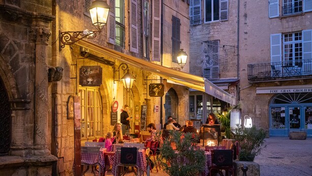 Un café al aire libre con mesas y sillas, vista a la calle y un edificio con un letrero.