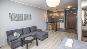 Sala moderna com sofá cinza, uma mesinha e uma cama com toalha branca dobrada. Há uma cozinha com um banco alto e uma porta de madeira.