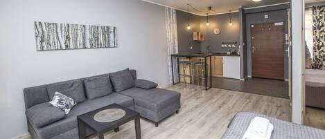 Una sala moderna con un sofá gris, una mesita y una cama con una toalla blanca doblada. Hay una zona de cocina con un taburete alto y una puerta de madera.