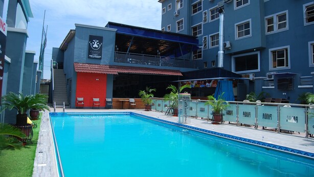 A swimming pool with a red building in the background, surrounded by potted plants and a fence. - DAWN CARRINGTON HOTELS AND SUITES (Lagos)