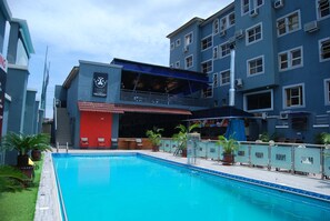 A swimming pool with a red building in the background, surrounded by potted plants and a fence. - DAWN CARRINGTON HOTELS AND SUITES (Lagos)